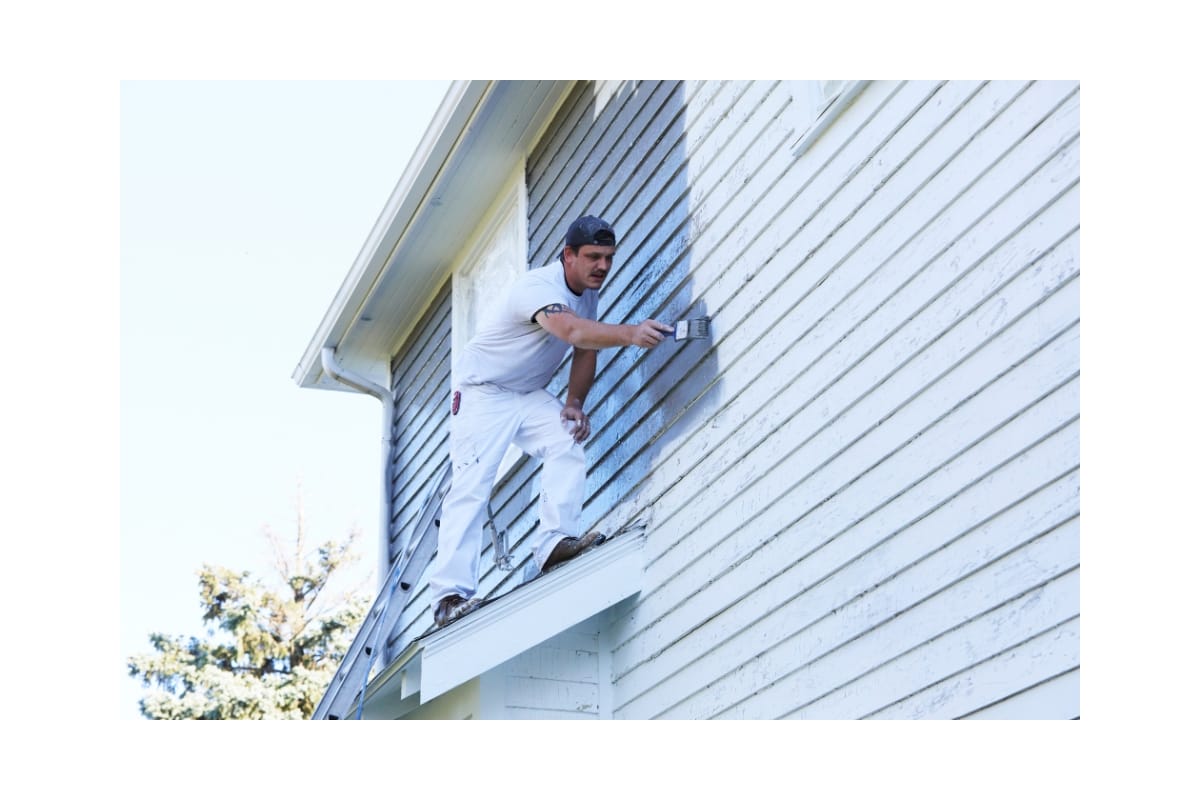 A residential contractor in white overalls is providing residential painting services to the exterior of a house from a high ladder, carefully applying paint to the side paneling with a brush.