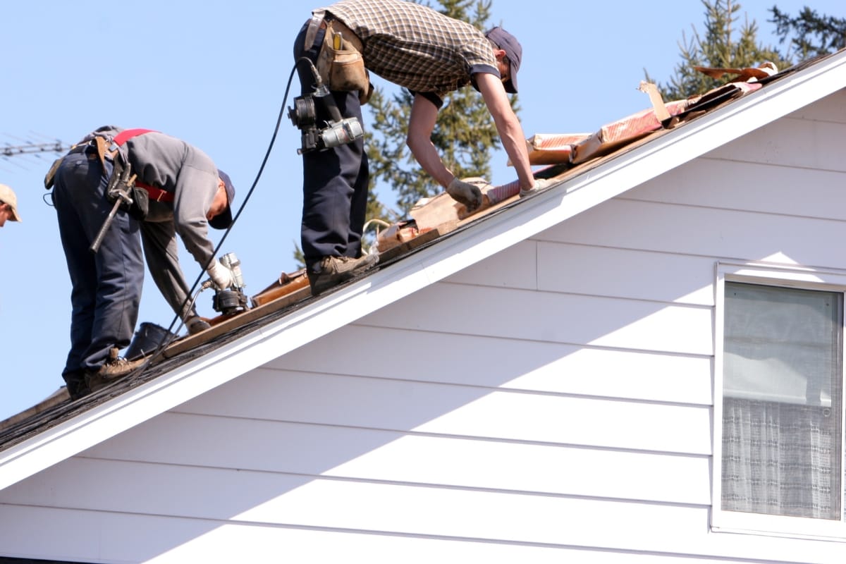 Two residential contractors are repairing a sloped roof on a white house, using tools and materials.