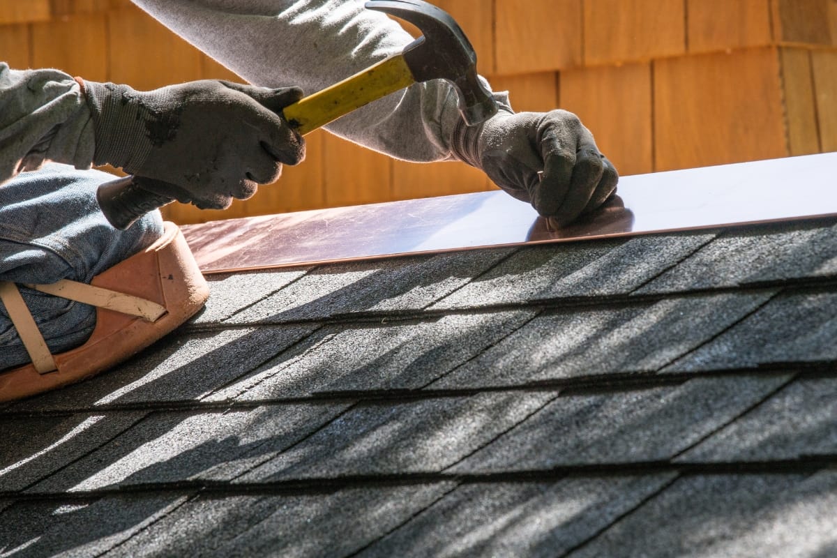 A person wearing gloves uses a hammer to secure metal flashing on the edge of a shingled roof, showcasing a skill often seen in residential contractors.