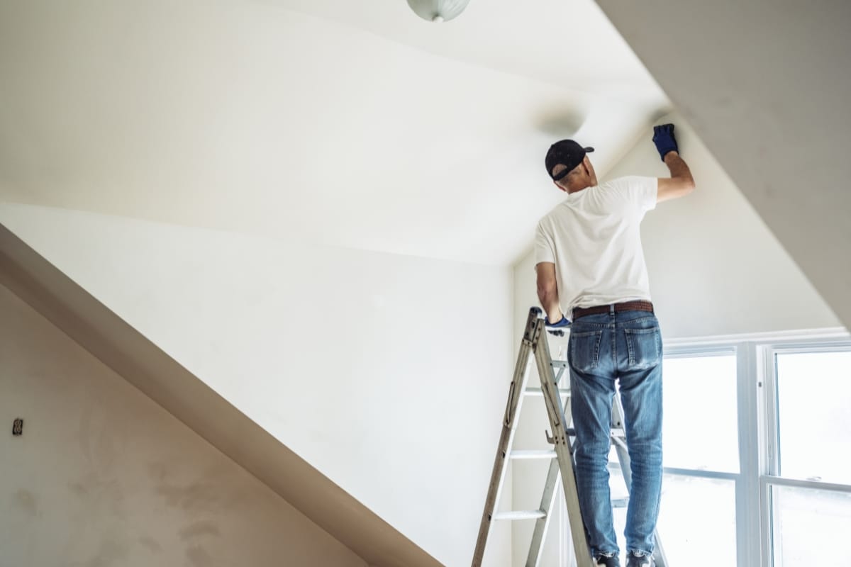 A person wearing a cap and gloves stands on a ladder painting a sloped ceiling in a white room, illuminated by sunlight streaming through the window—a task that could easily be handled by experienced residential contractors.