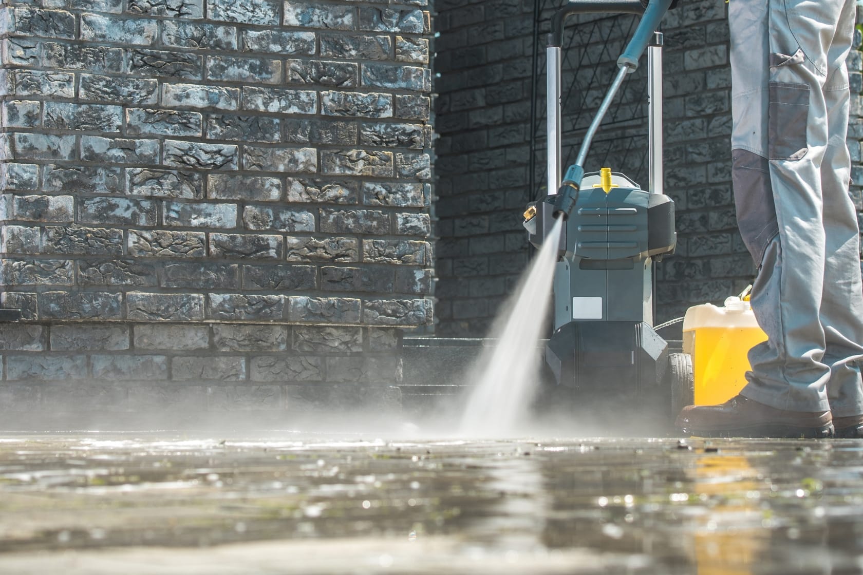 Person using a pressure washer to clean a stone patio in front of a brick wall.