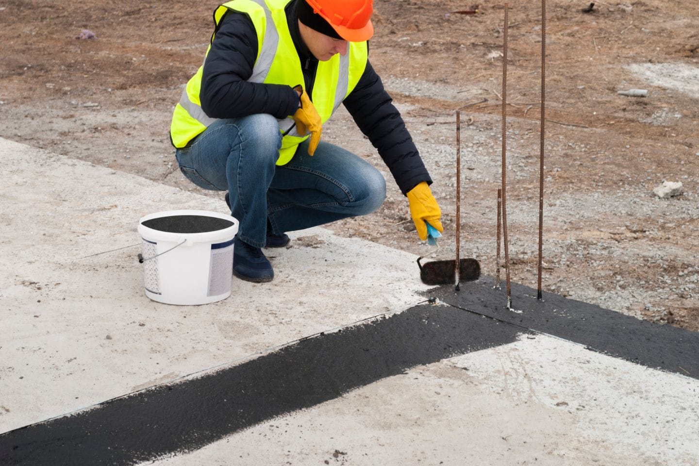 A person wearing a hard hat and high-visibility vest applies black waterproofing material to a concrete surface at a construction site, ensuring it's sealed for future use as part of a home foundation. A bucket of the material is nearby.