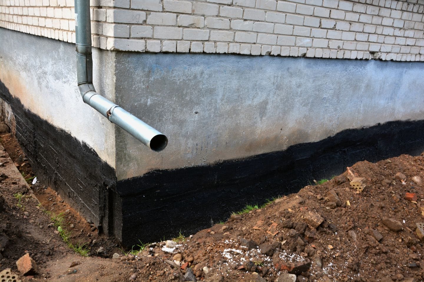 Close-up of a home's foundation being waterproofed with a black coating, while a downspout directs rainwater away from the base. Dug-up soil and debris are visible in the foreground.