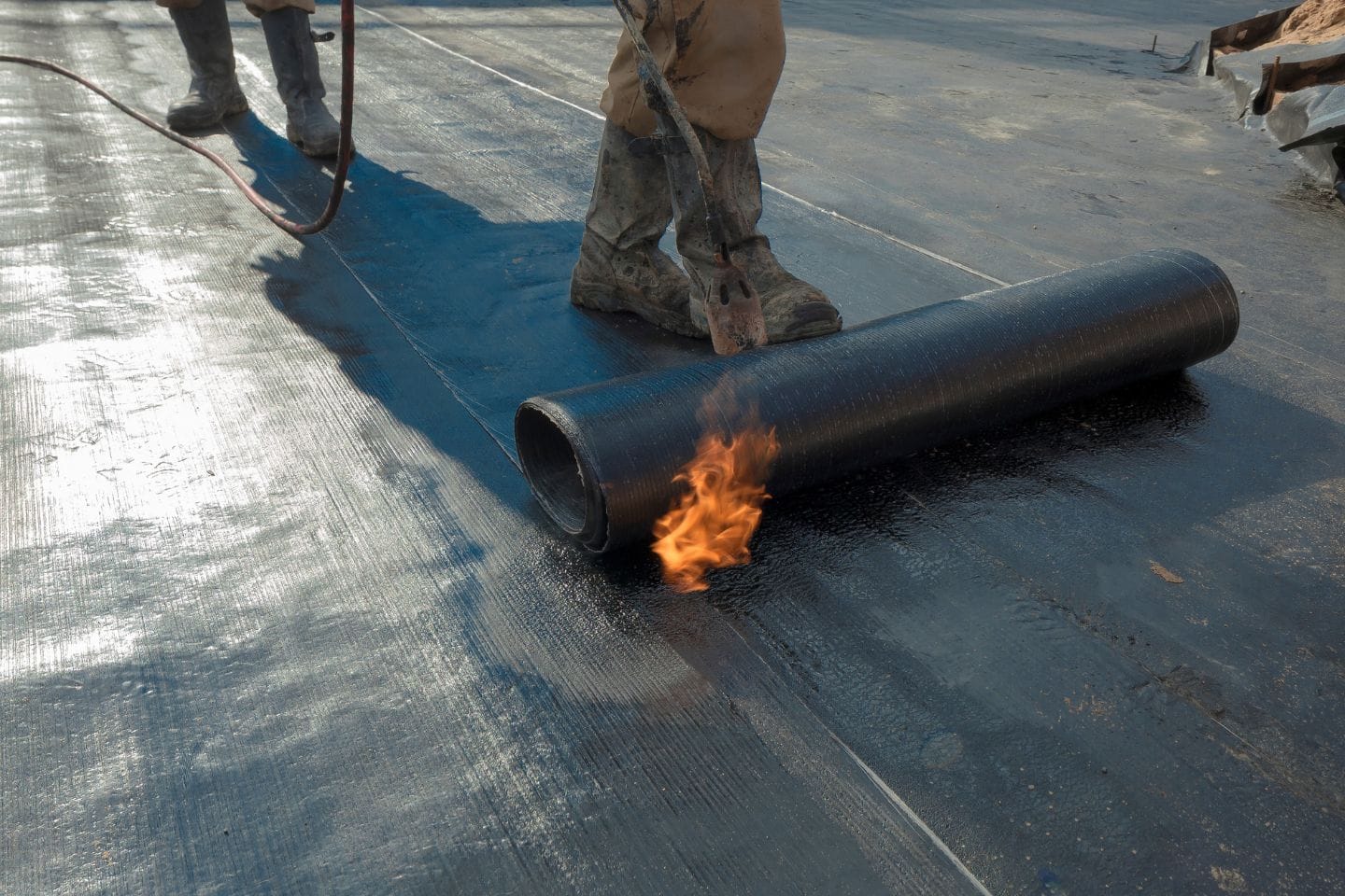 Workers using a blow torch to install rolled roofing material on a flat home surface.