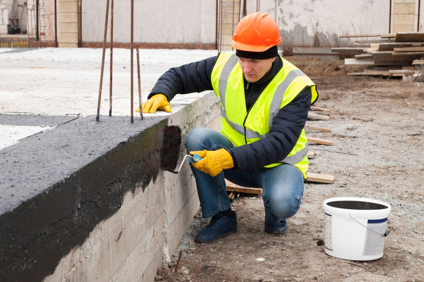 A worker in a high-visibility jacket and orange helmet applies a coating with a brush to a concrete structure at what will soon be someone's home. A white bucket is placed nearby.