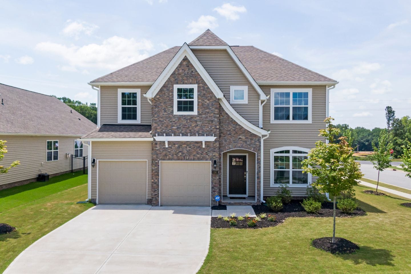 A charming two-story home with beige siding and stone accents, featuring a double garage and a well-maintained front yard, situated in a suburban neighborhood.