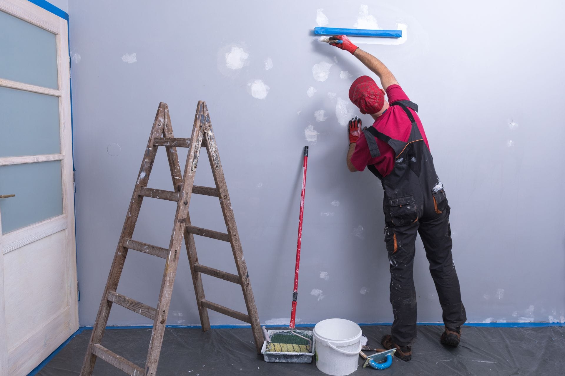 A person in work clothes paints a wall light blue using a roller while standing on the floor, turning their home into a serene haven. A ladder, paint bucket, and paint tray are nearby.