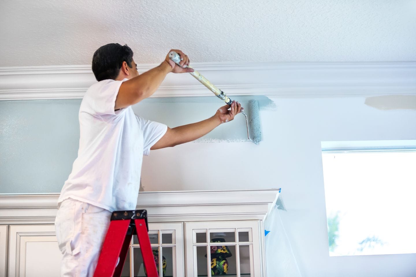 A person stands on a ladder painting a wall blue near the ceiling in their home.