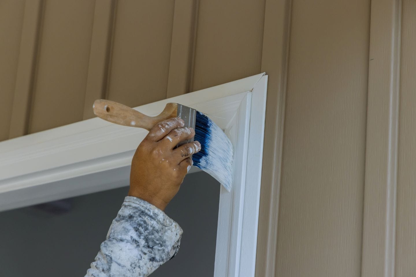 A person is painting the trim of a door with a brush, applying white paint to the frame. The background features beige siding, capturing a serene home improvement scene.