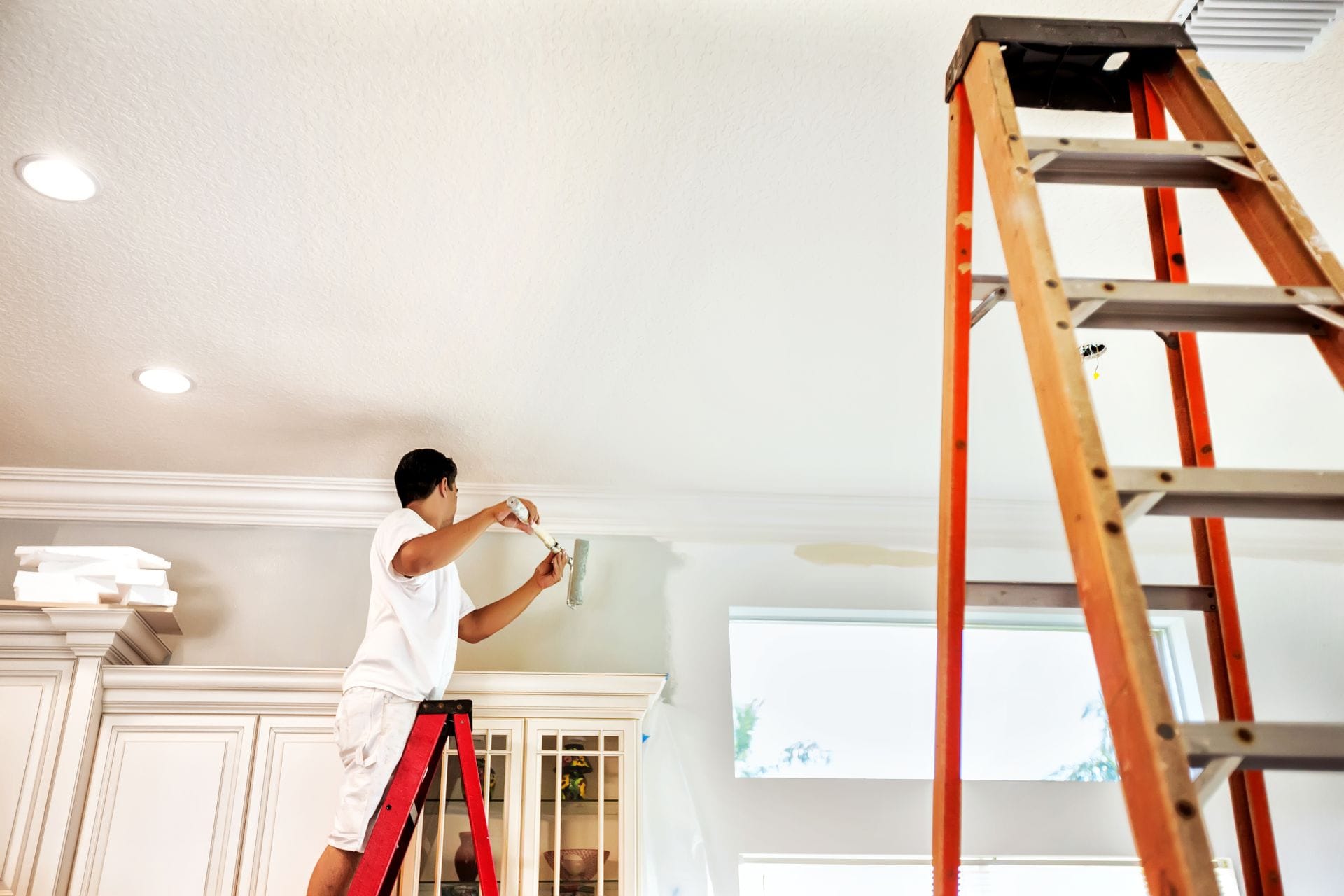 A person stands on a ladder, painting the upper wall near the ceiling in a bright home, with another ladder nearby.