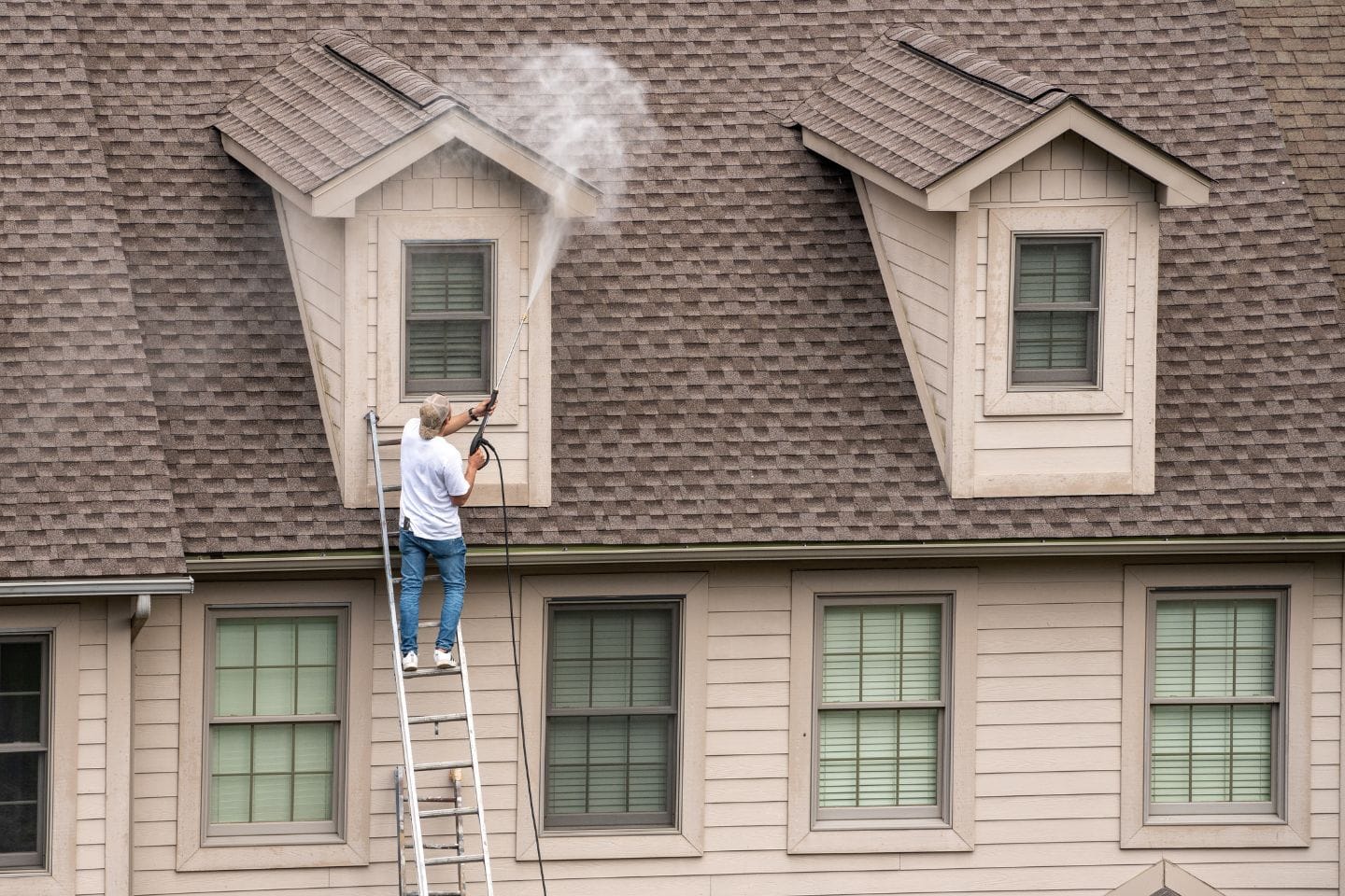 A person on a ladder is pressure washing a home's shingled roof, focusing on an area around a dormer window.