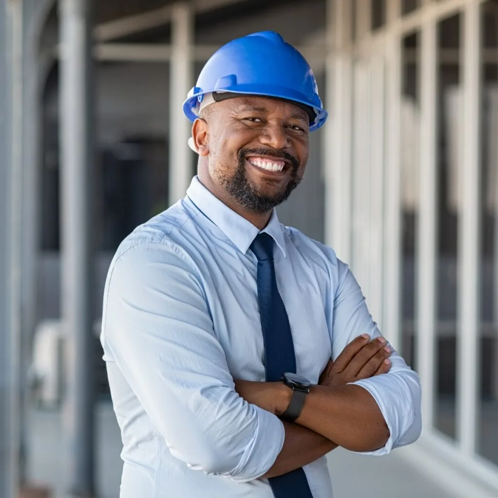 Person in a blue hard hat and shirt, smiling confidently with arms crossed, stands proudly in front of an innovative building.