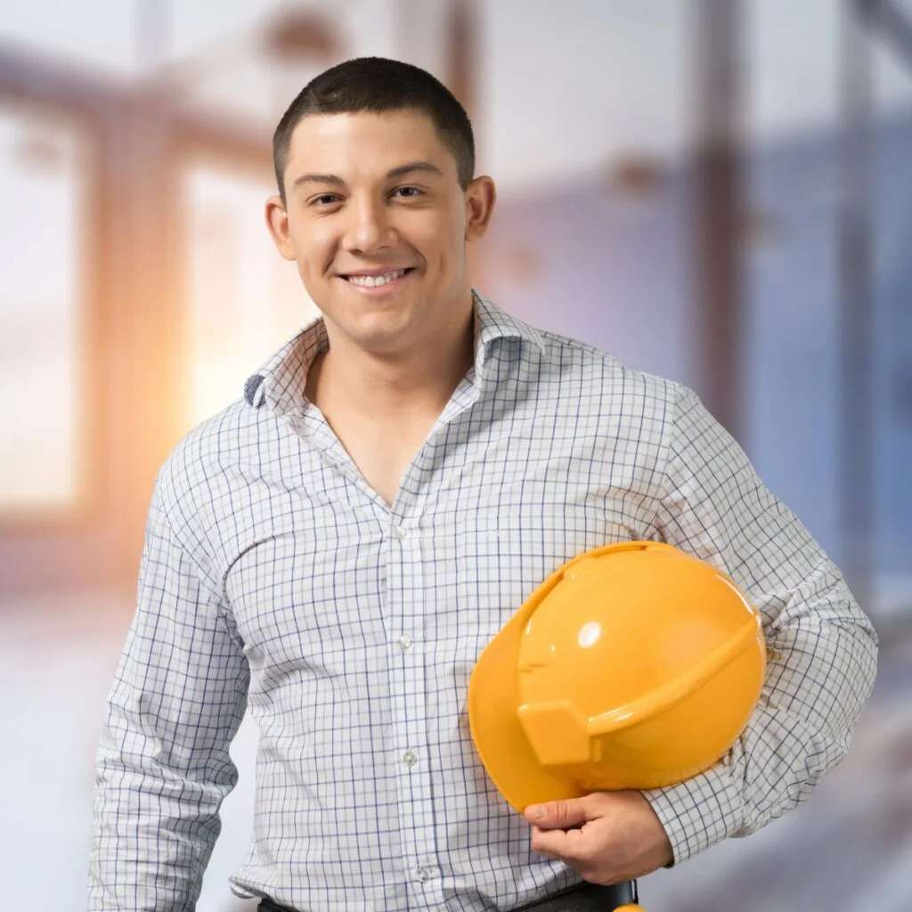 A person in a checkered shirt smiles confidently while holding a yellow hard hat in a brightly lit room, radiating the spirit of determination and professionalism.