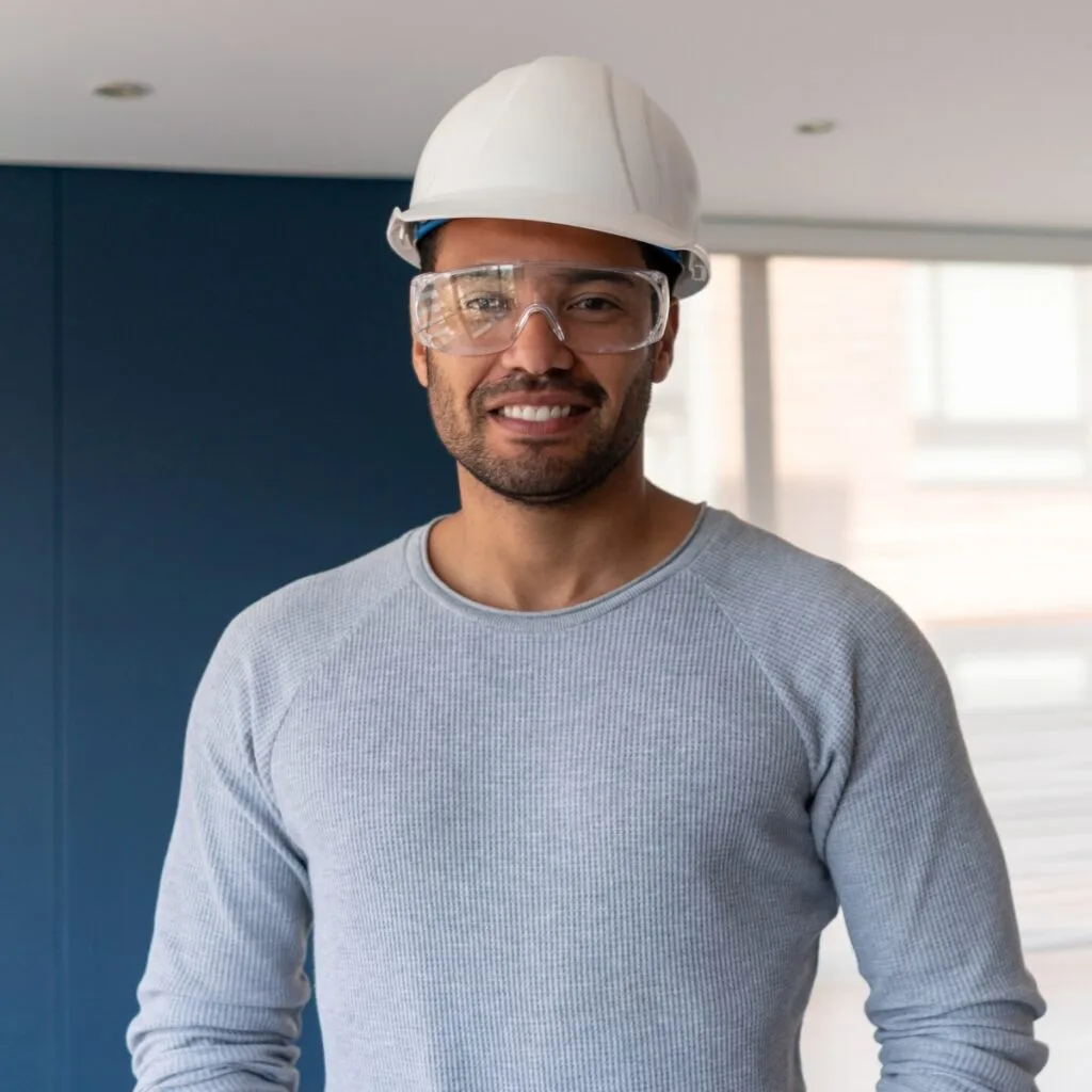 A diligent worker, clad in a white hard hat and safety goggles, sports a cozy gray sweater within an industrial setting. I'm sorry if more specifics are needed, but the scene showcases robust safety practices in action amidst the bustling indoors.