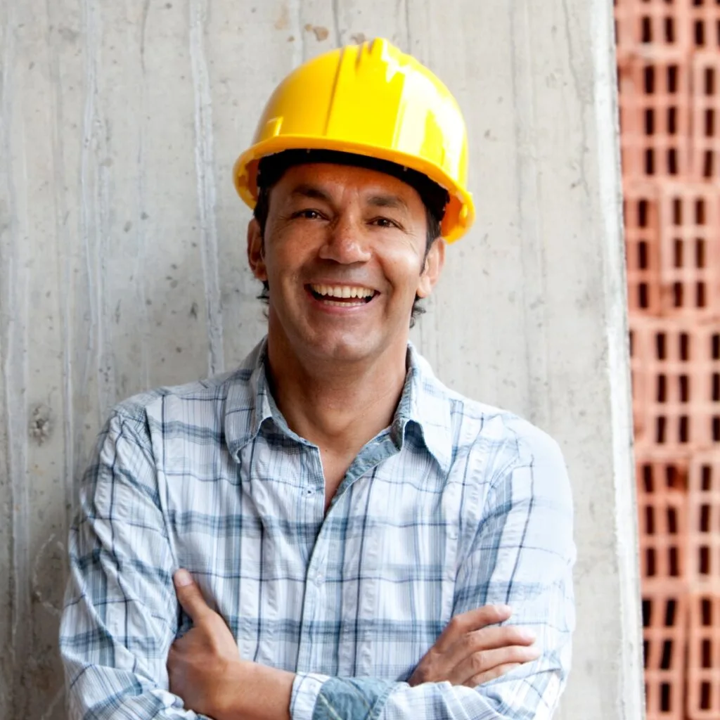 About to start his next big project, a man in a plaid shirt and yellow hard hat smiles with arms crossed, standing confidently against a concrete wall.