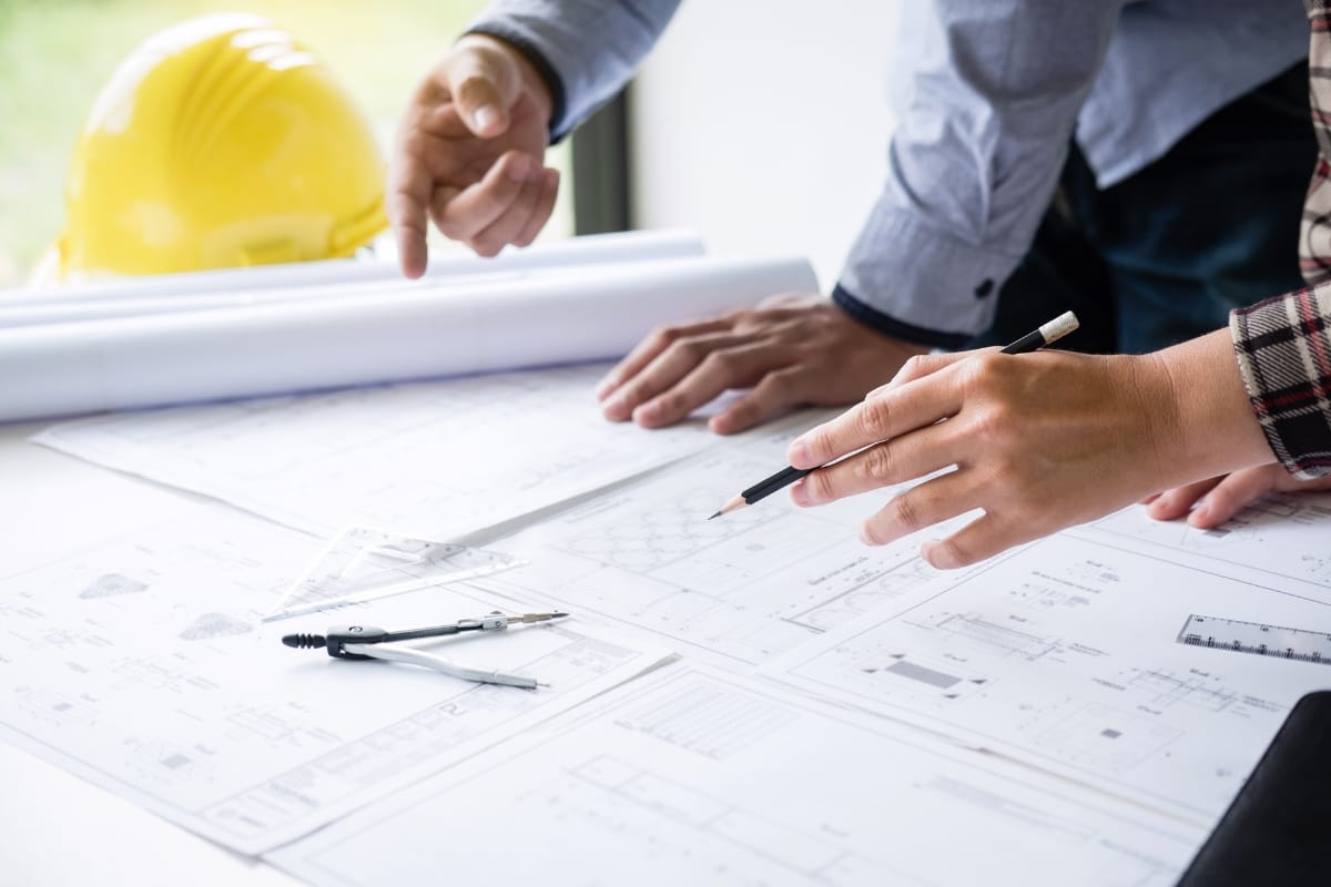 Two people are discussing architectural blueprints on a table, equipped with a pencil and compass, emphasizing their professional services. A yellow hard hat sits in the background, symbolizing their commitment to safety and precision.