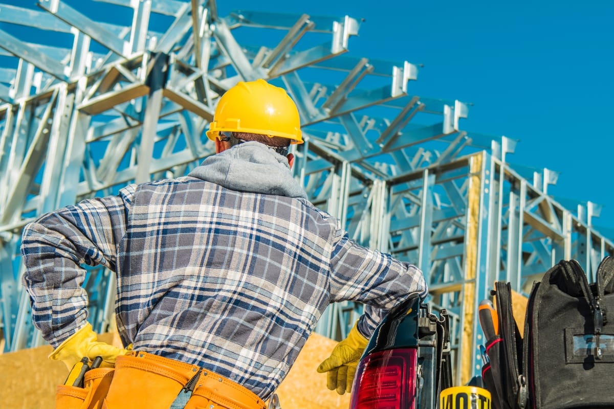 A construction worker in a hard hat and plaid shirt services the building frame under a clear blue sky, with equipment bags visible nearby.