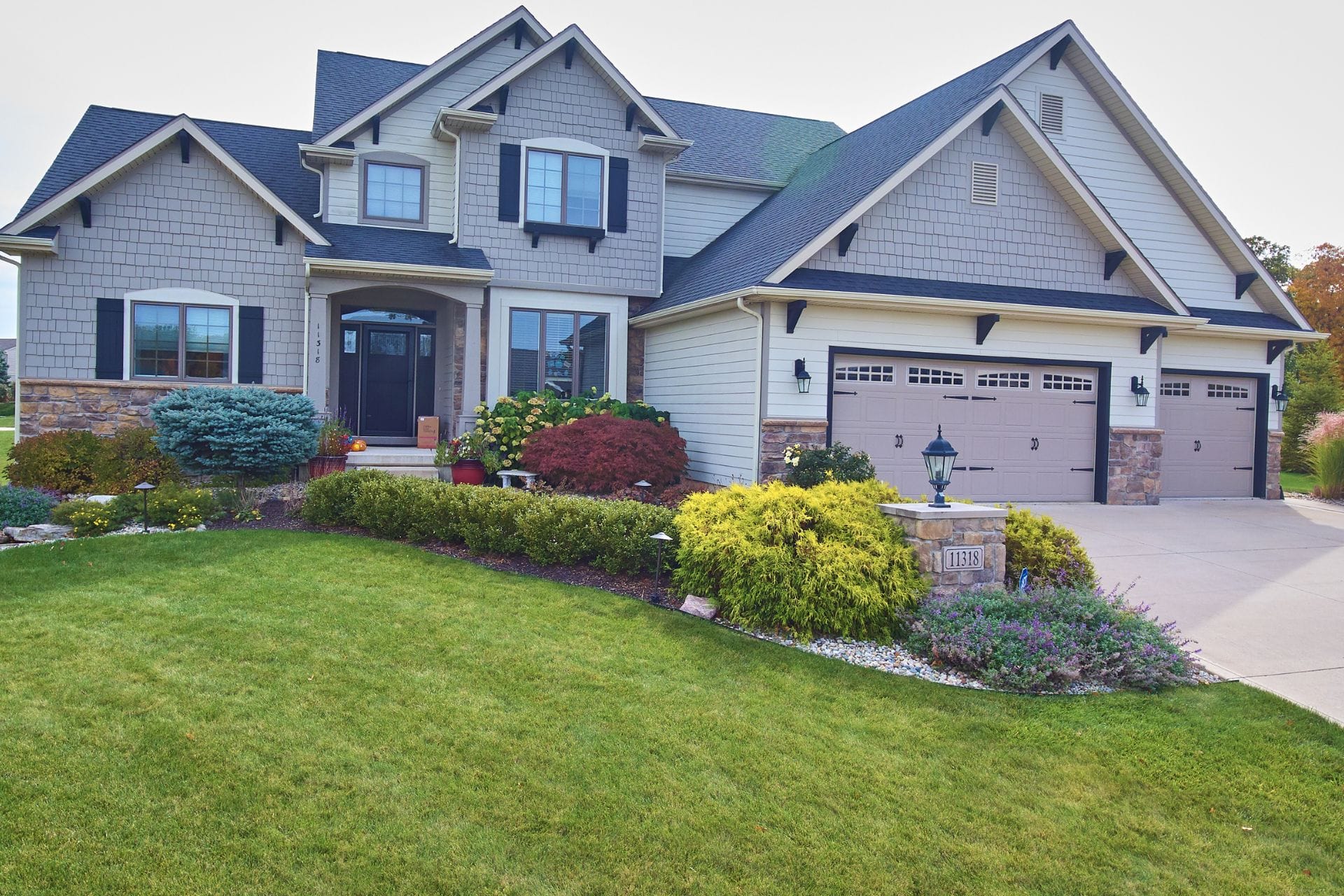 A modern suburban house with gray siding, black shutters, and a three-car garage showcases exquisite exterior painting. It's surrounded by well-maintained landscaping and a green lawn.