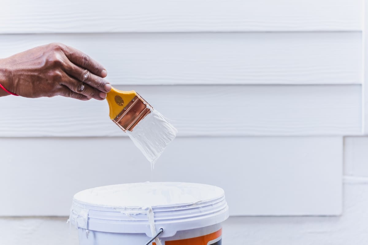 A hand holds a paintbrush dripping white paint over a paint bucket, perfect for exterior painting. White siding is visible in the background, capturing the home's transformation.