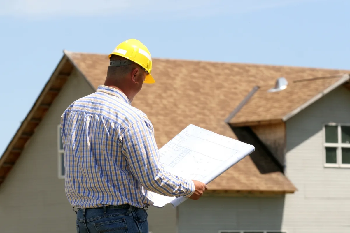 A person wearing a yellow hard hat holds blueprints, standing confidently in front of a house under construction, contemplating the design intricacies.