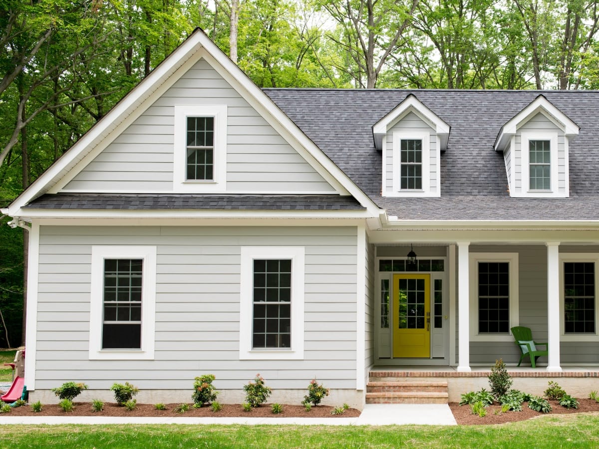 A modern gray house with white trim and expert roofing features two dormer windows, a porch with white columns, and a bright yellow front door, surrounded by trees and landscaping.