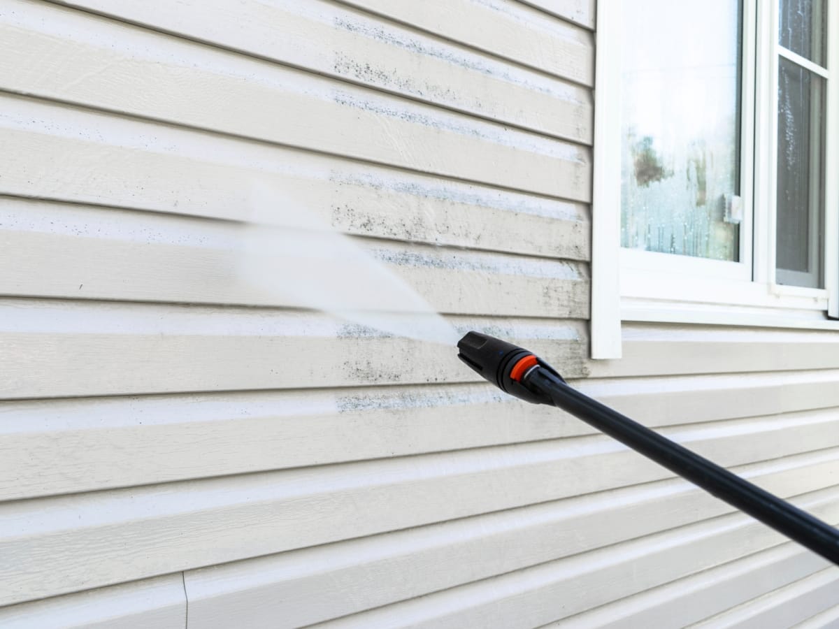 A power washer is expertly spraying water onto a house's white siding, efficiently cleaning off dirt and grime near a window. This pressure washing service ensures the exterior looks pristine and well-maintained.