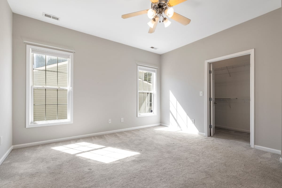 This spacious room features a beige carpet, light gray walls, ceiling fan, two windows, and a walk-in closet. Designed for comfort and versatility, it's perfect for relaxing or setting up your home office services.