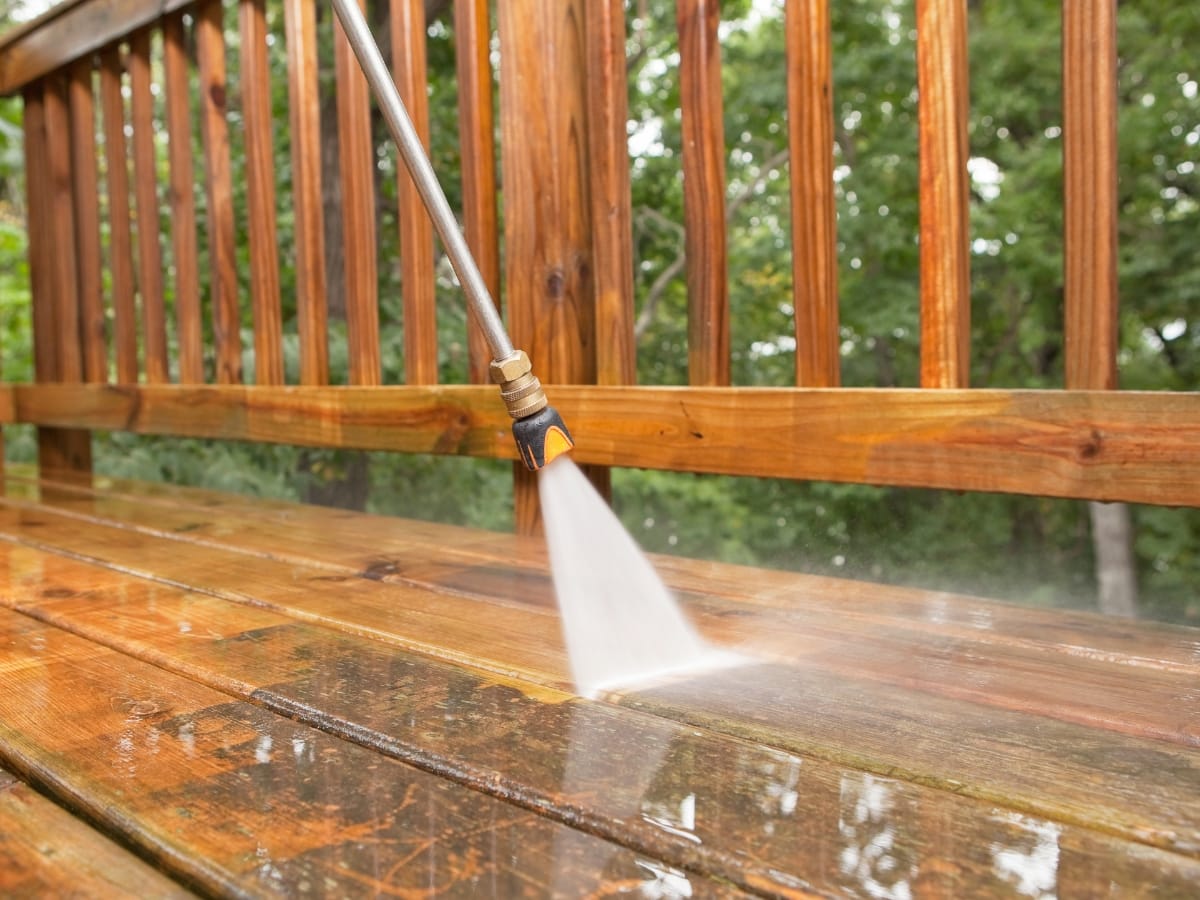 A power washer performs exterior maintenance by cleaning a wooden deck with water jets in a forested area.