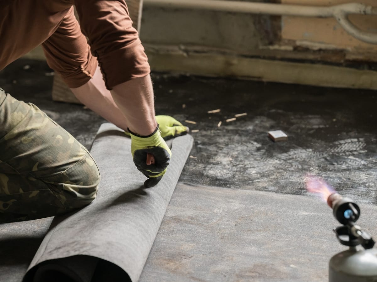 A person in gloves kneels while installing a roll of waterproofing material on a commercial floor. A blow torch is seen in the foreground.