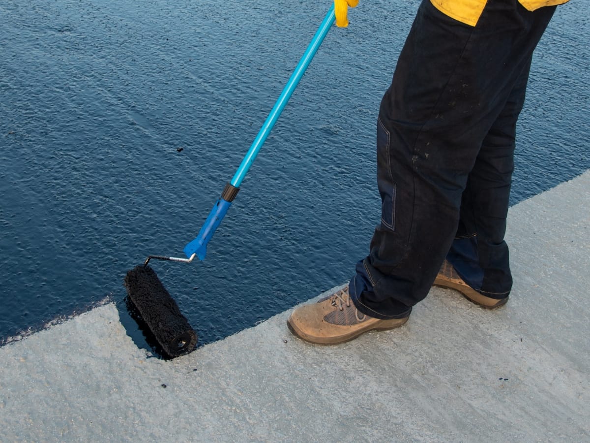 A person expertly applies black waterproofing paint with a roller, ensuring the flat surface of the commercial building is well-protected.