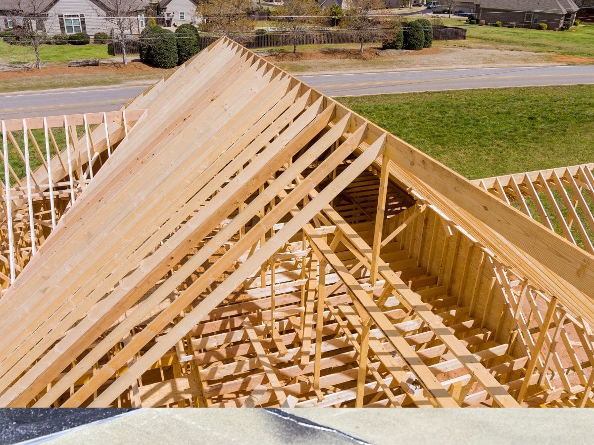 Residential wooden framework of a house under construction, showcasing roof trusses and beams against a suburban neighborhood backdrop, is part of a storm restoration project.